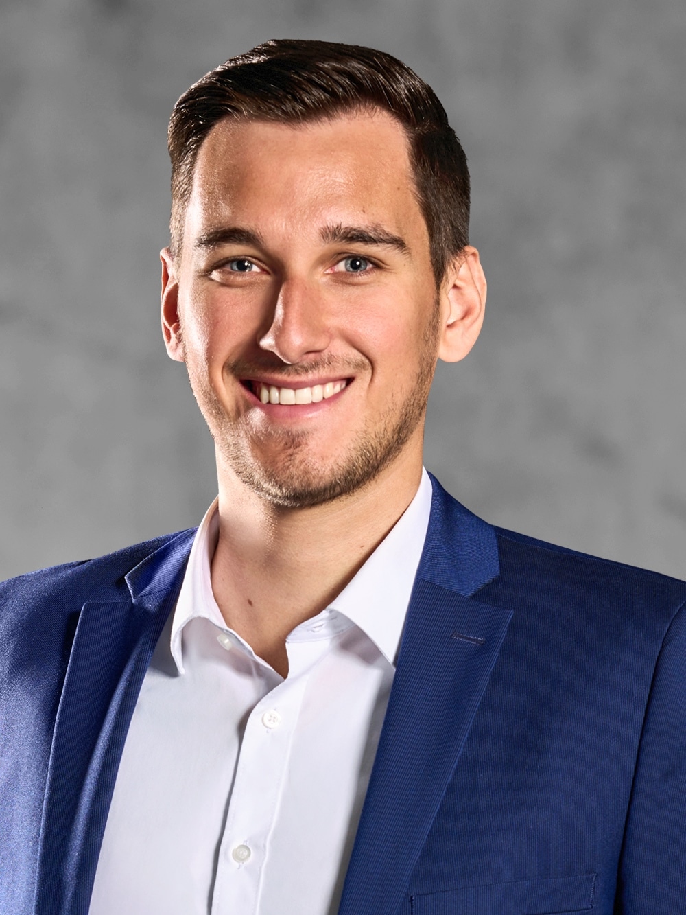Portrait von Jannis Adam. Lächelnder junger Mann mit braunem Kurzhaarschnitt trägt ein weißes Hemd und ein blaues Jackett.