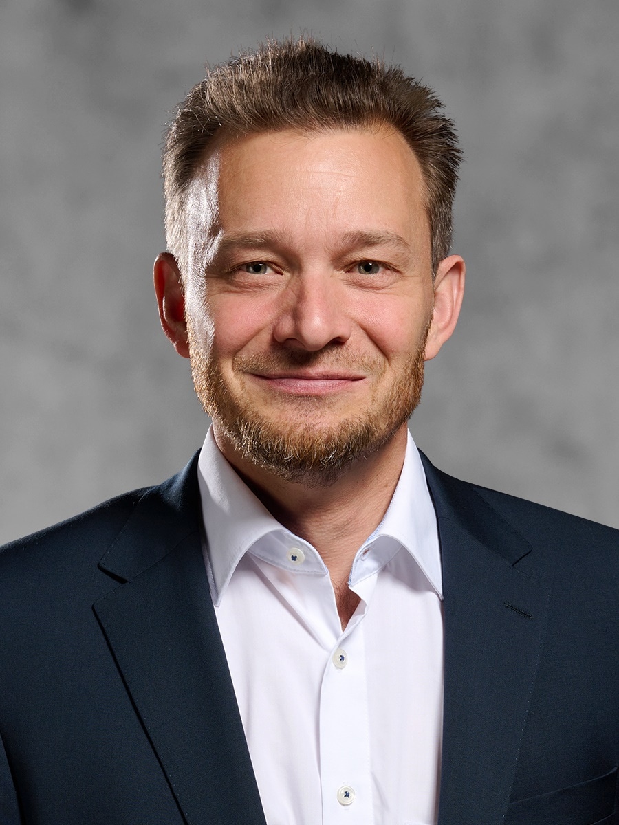 Portrait von Stefan Duschl. Lächelnder Mann mit braunem Haar und Bart. Er trägt ein weißes Hemd und schwarzes Jackett. 