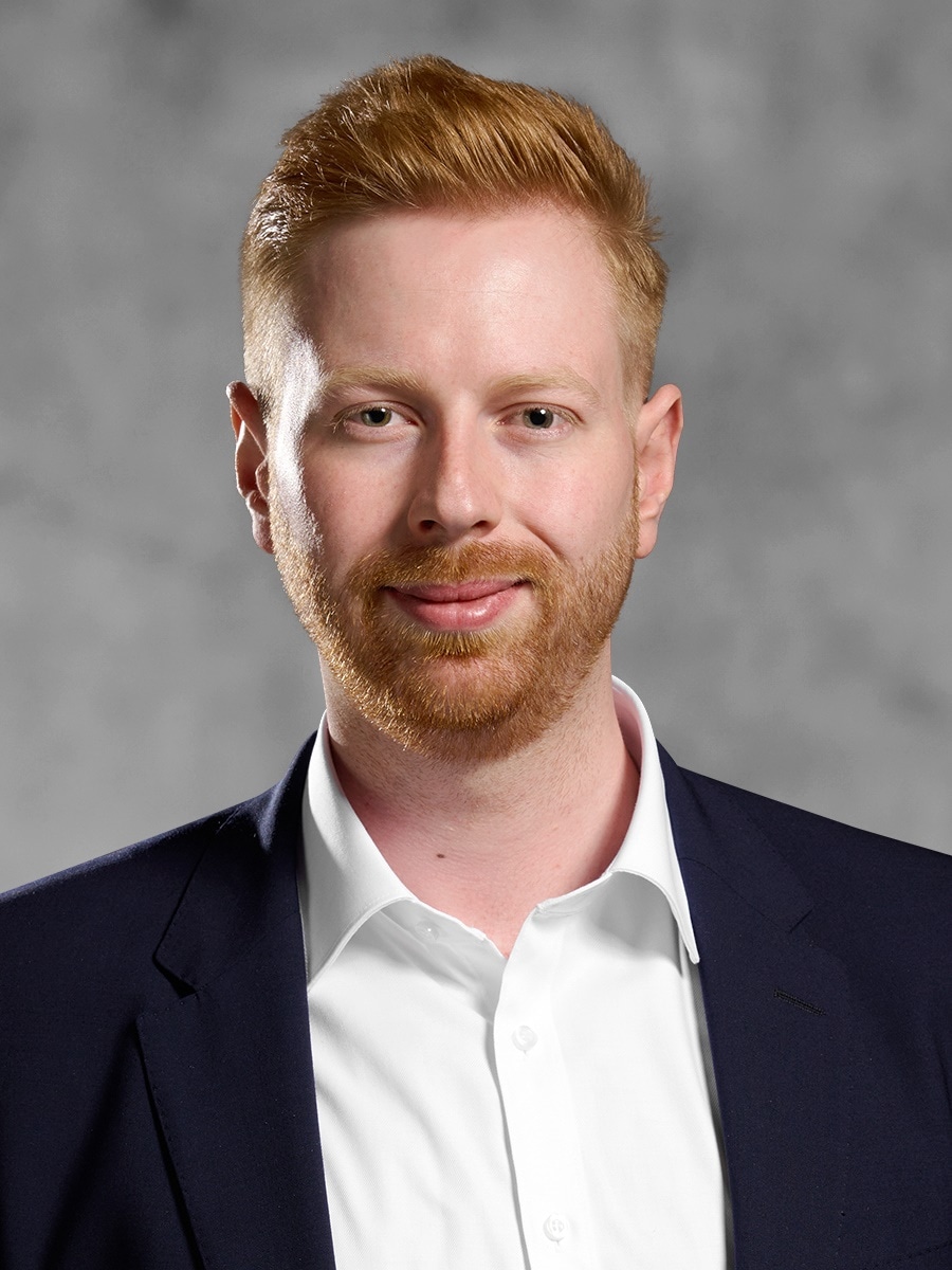 Portrait von Marius Harsch. Rothaariger Mann mit Kurzhaarschnitt und einem kurzen Vollbart in weißem Hemd und Jackett. 
