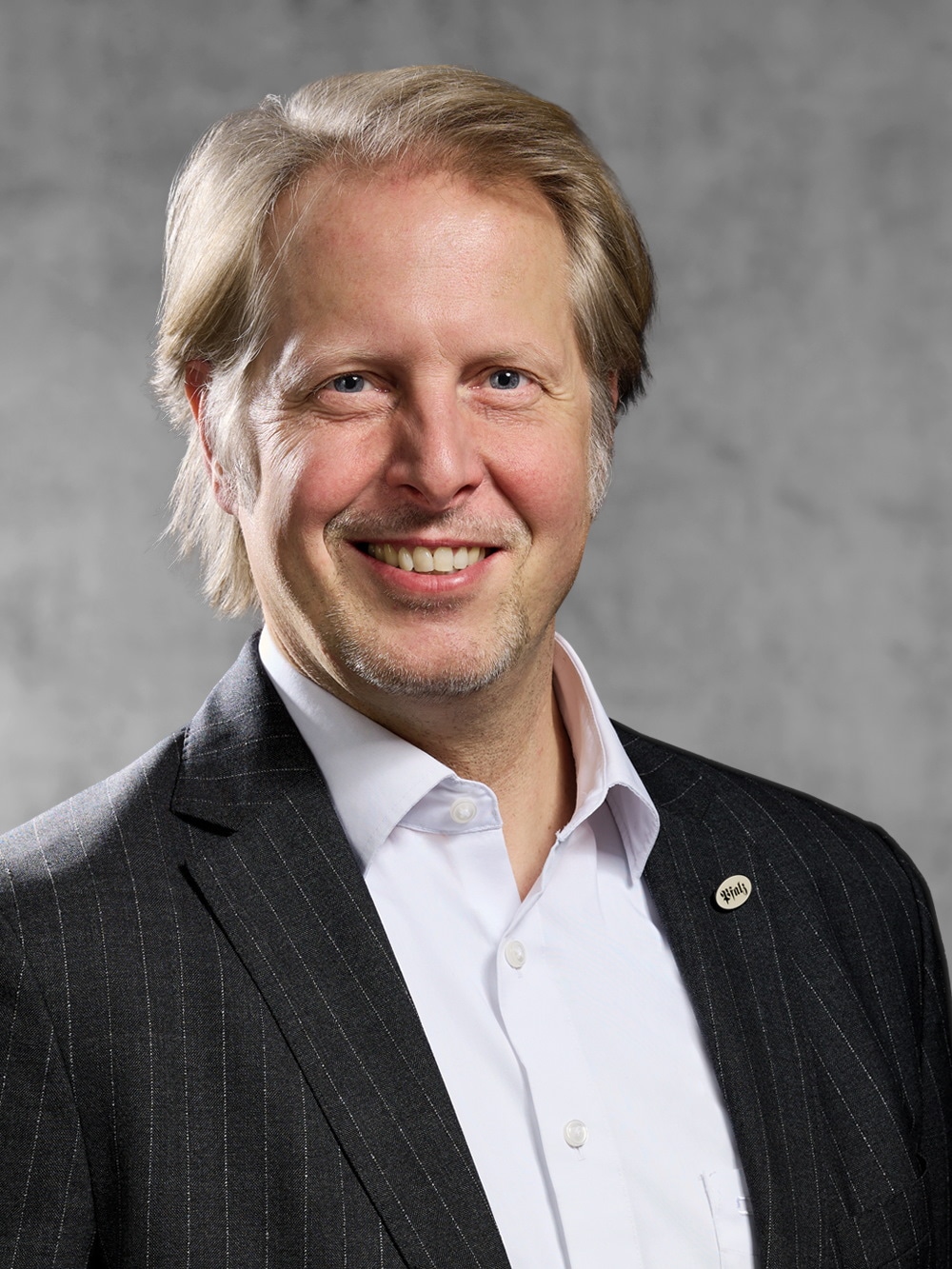 Portrait von Axel Herrle. Mann mit graublondem Haar und 3-Tagesbart in weißem Hemd und Jackett mit Nadelstreifen. 