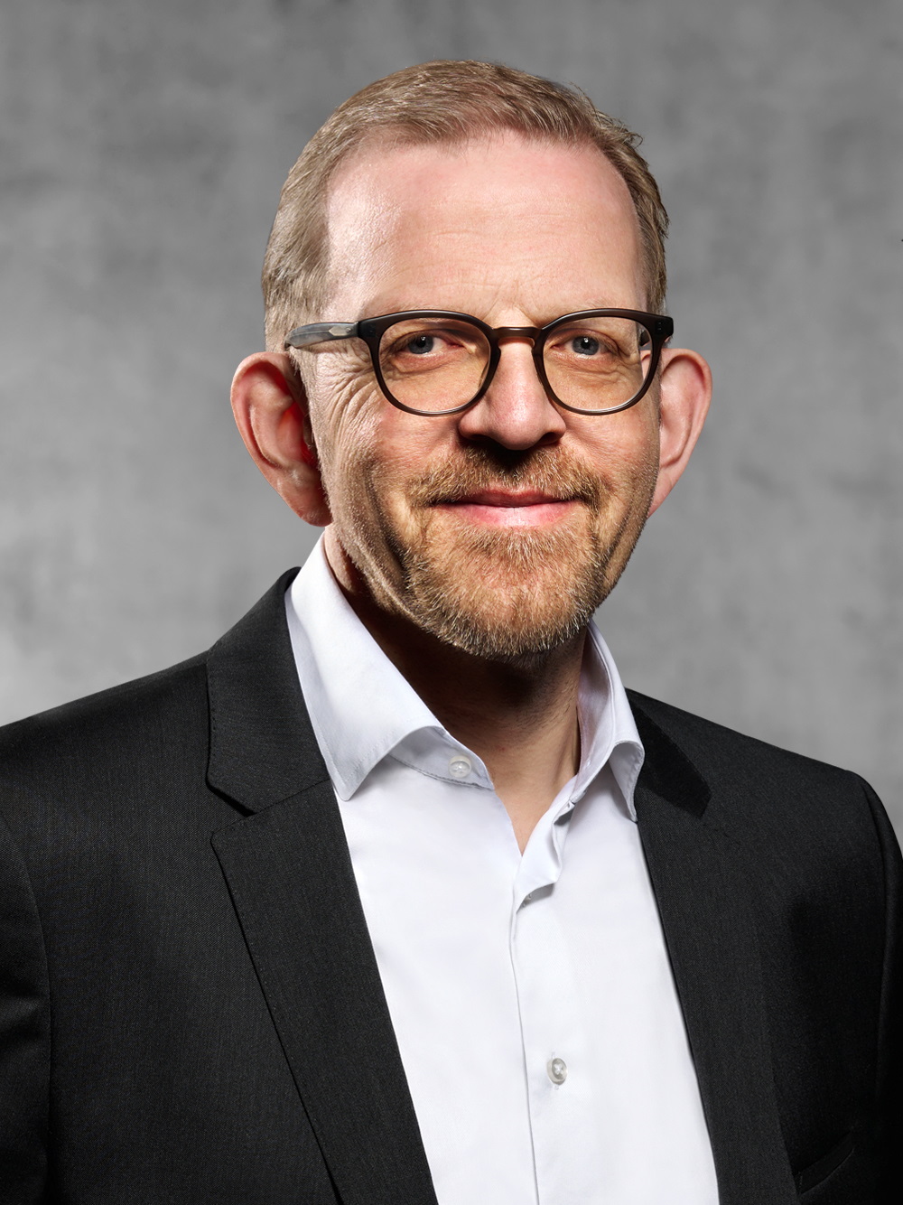 Portrait von Michael Kleerbaum. Mann mit blondem Kurzhaarschnitt und kurzen Bart, trägt Brille, weißes Hemd und Jackett