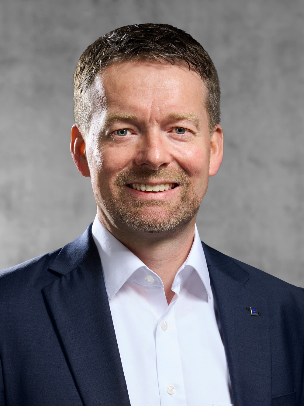 Portrait von Manfred Kratz. Mann mit kurzem dunklen Haar und kurzem Bart, trägt weißes Hemd und blaues Jackett.