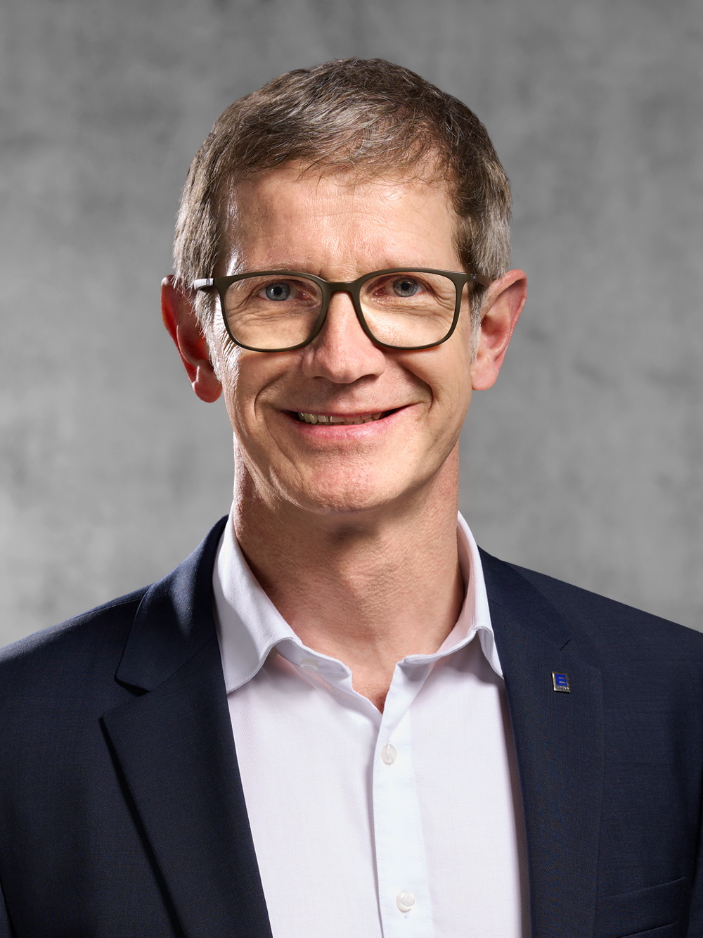 Portrait von Matthias Lein. Mann mit Brille, rasiert, mit kurzem blondem Haar trägt ein weißes Hemd und ein blaues Jackett.