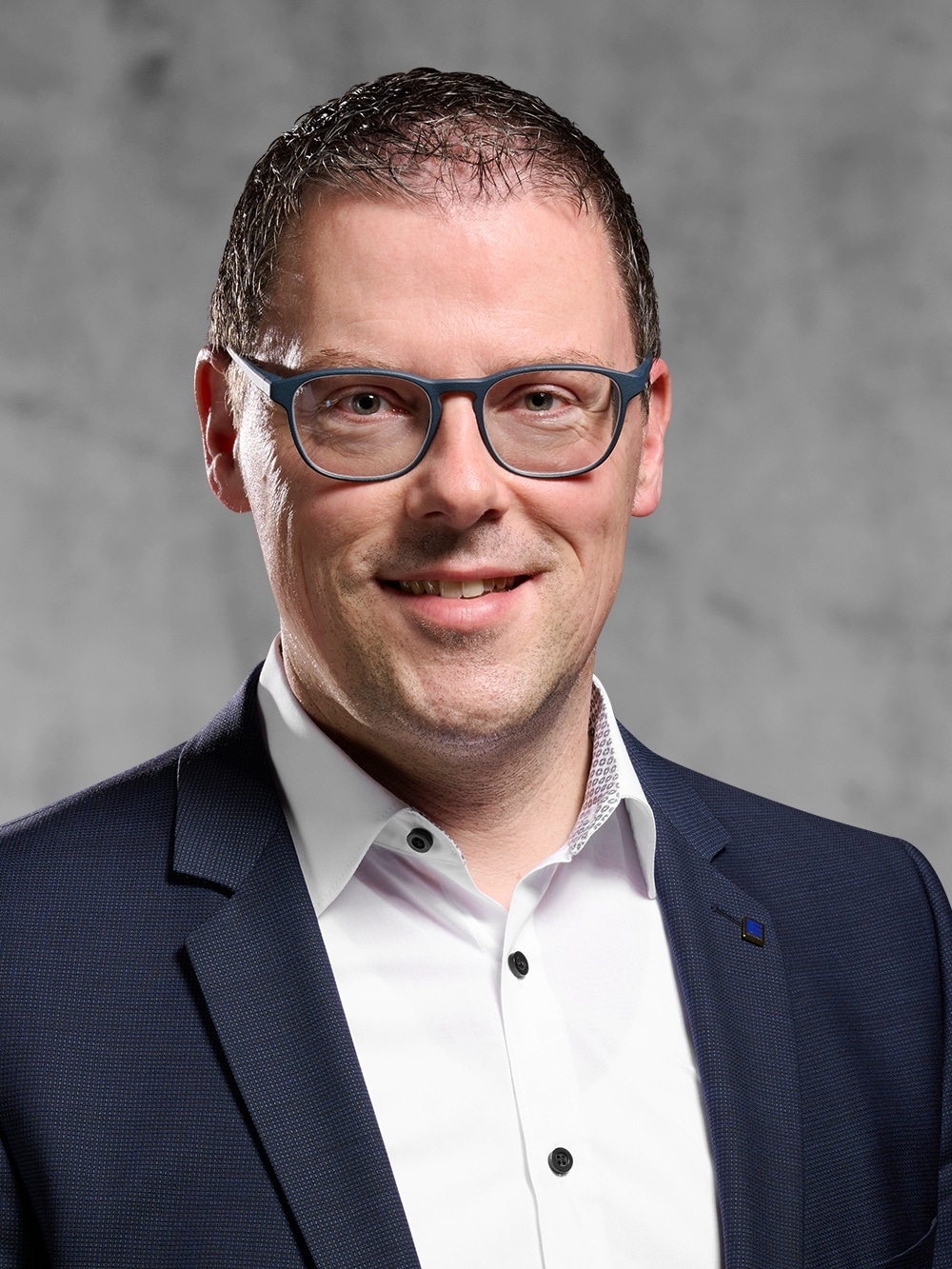 Portrait von Frank Meng. Mann mit Brille, Tagesbart, kurzem dunklem Haar, trägt ein weißes Hemd und ein blaues Jackett.
