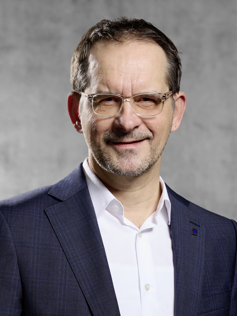 Portrait von Wolfgang Seiler. Mann mit kurzem dunklen Haar und kurzem Bart, trägt Brille, weißes Hemd und blaues Jackett