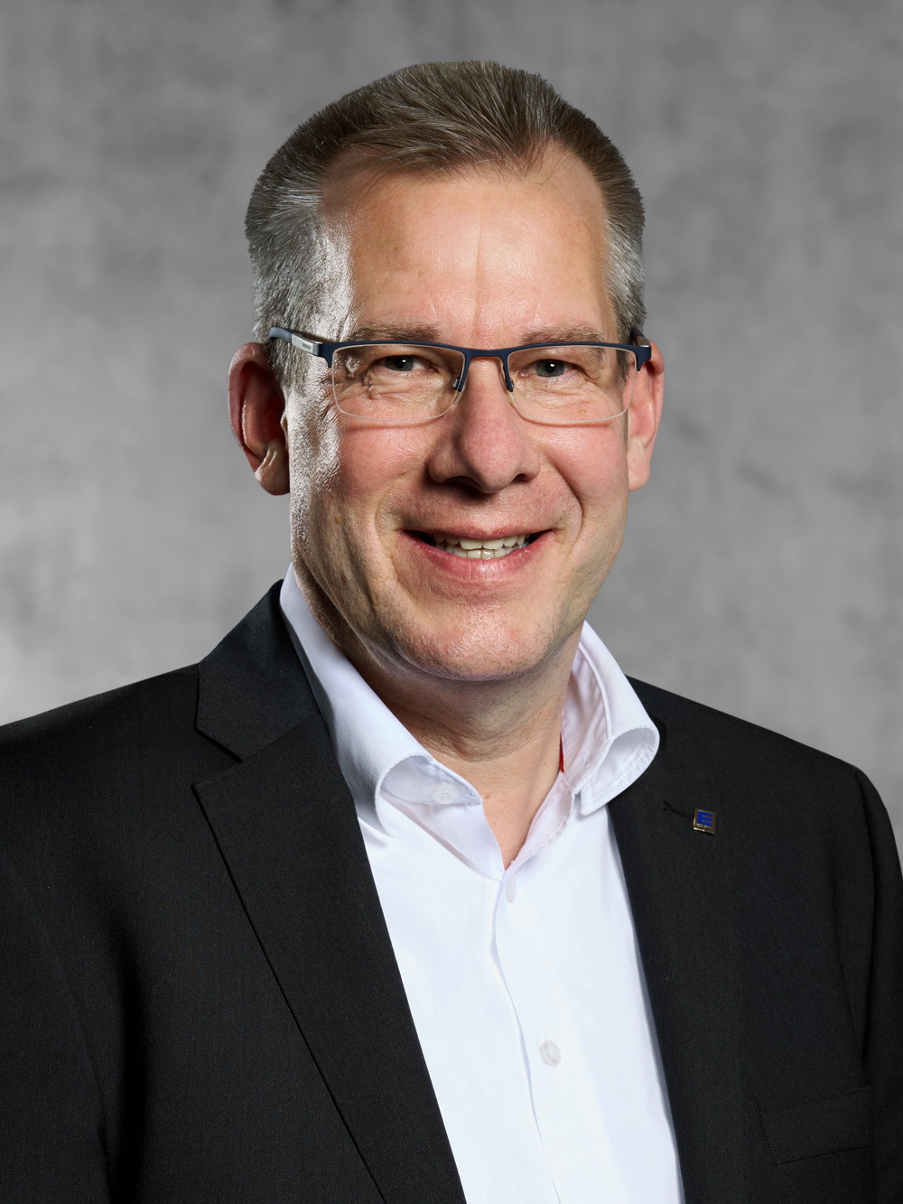 Portrait von Alex Weber. Mann mit kurzem graumeliertem Haar, lächelt, trägt Brille, weißes Hemd und schwarzes Jackett.