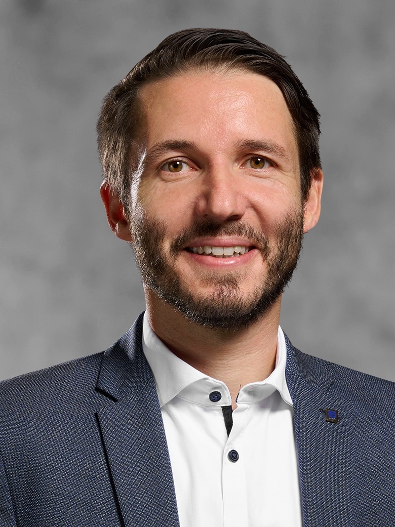 Portrait von Andreas Wilcek. Mann mit kurzem dunklem Haar und Bart, trägt ein weißes Hemd und ein graues Jackett.