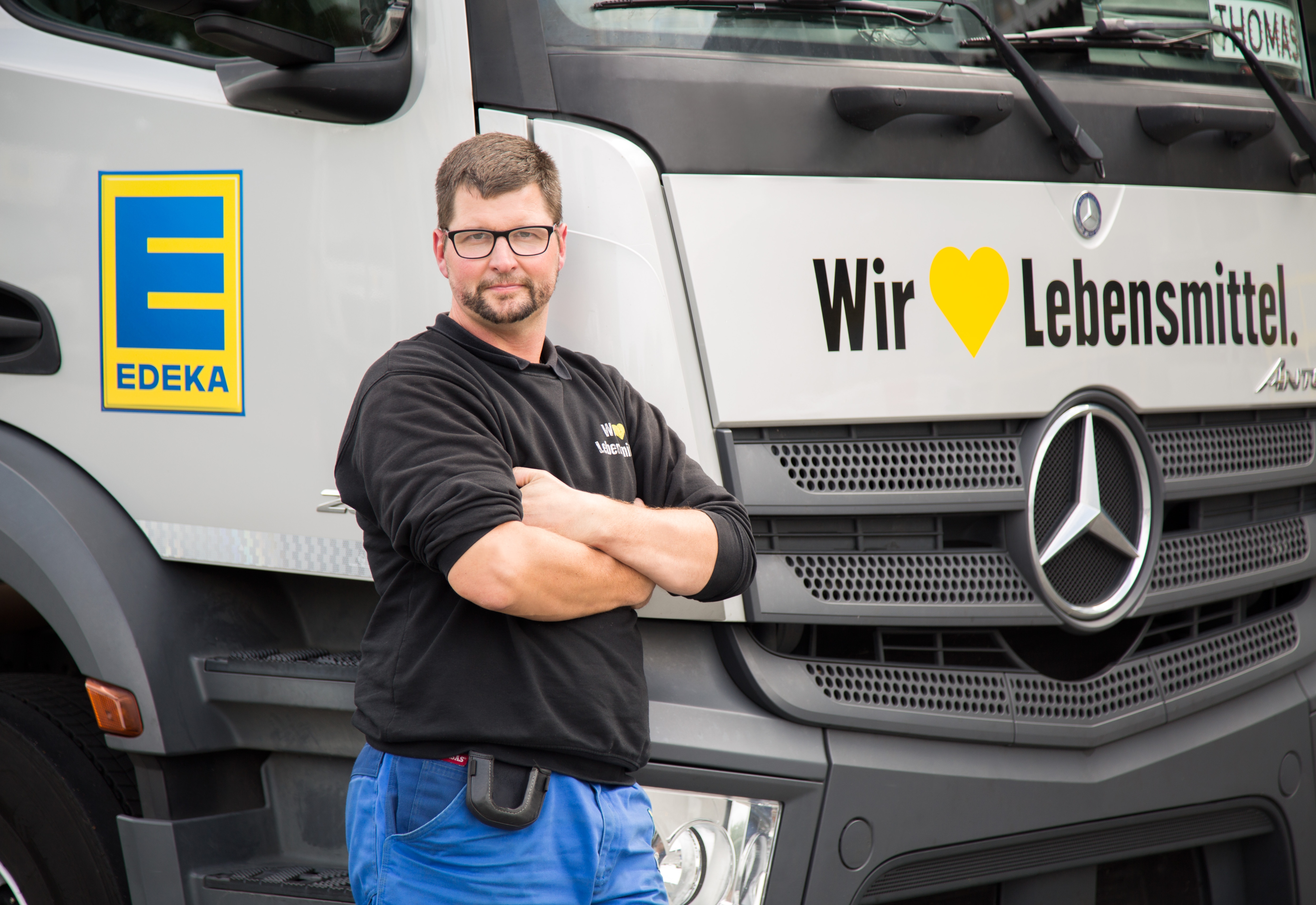 Mann mit Brille und dunklem Bart steht mit verschränkten Armen vor einem LKW mit der Aufschrift Wir lieben Lebensmittel.