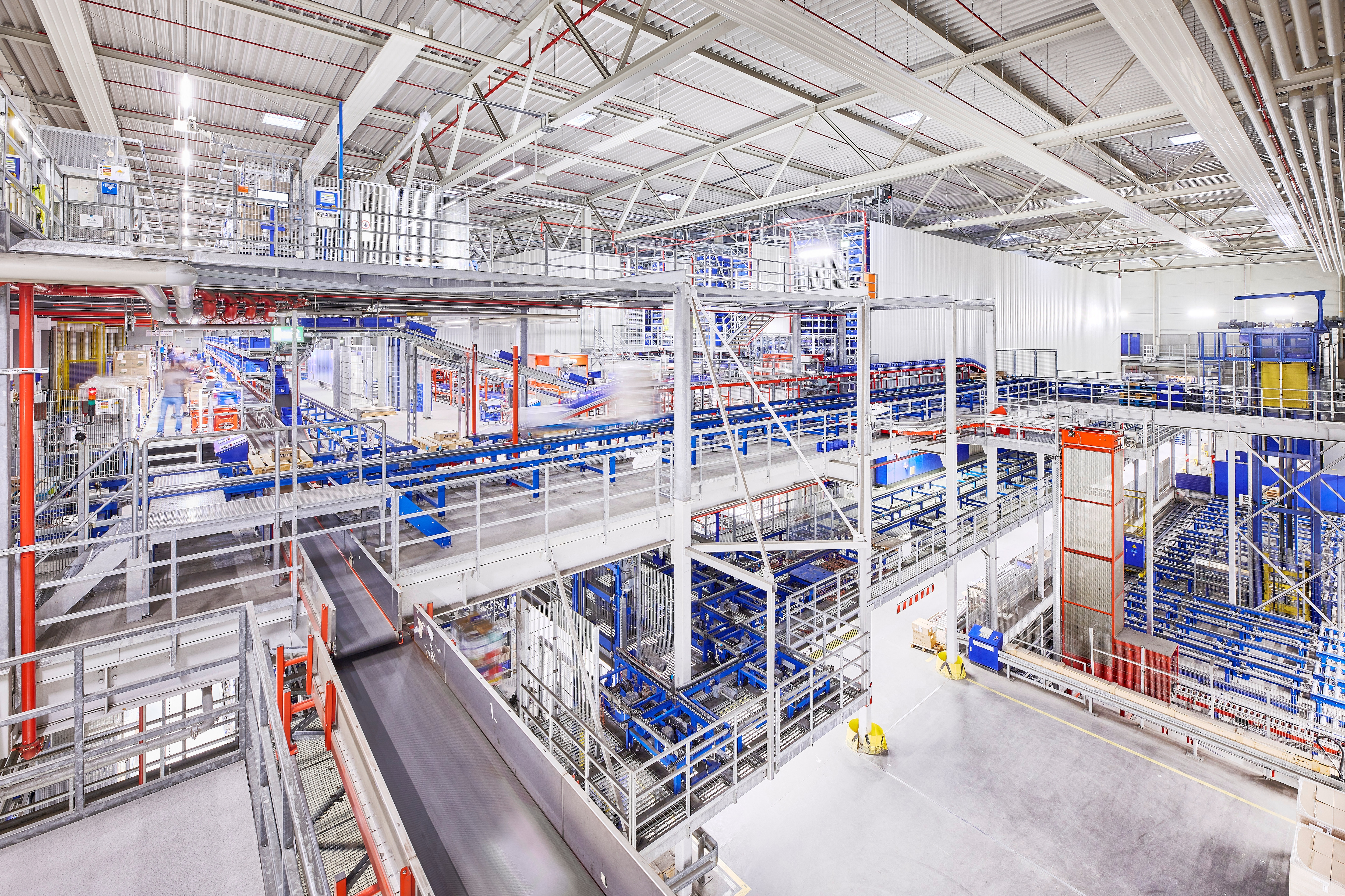 hell beleuchtetes Logistikzentrum mit mehreren Ebenen von Förderbändern und automatisierten Sortieranlagen.