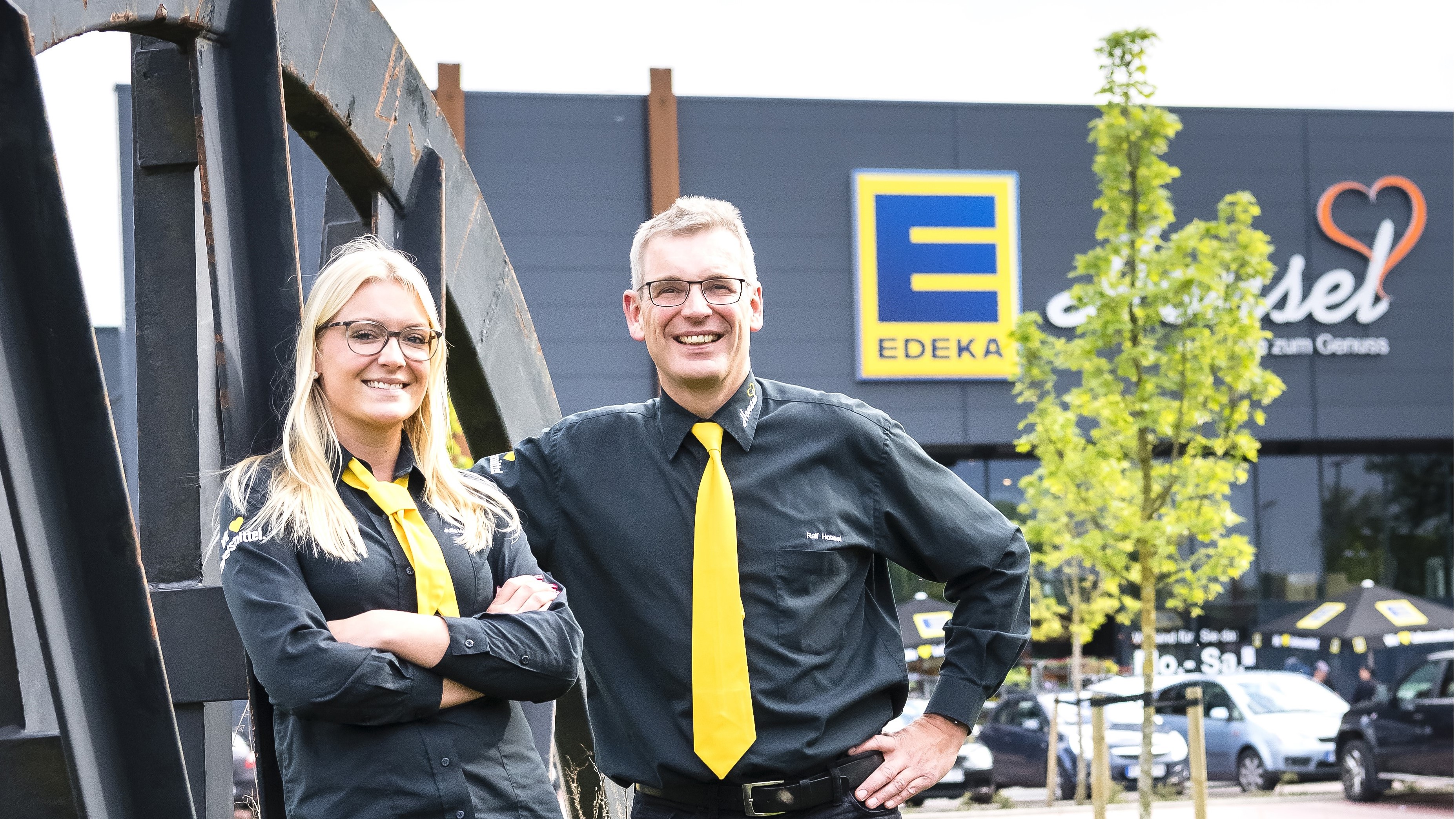 Eine Frau und ein Mann stehen vor einem großen EDEKA-Markt. Beide tragen schwarze Hemden von EDEKA und eine gelbe Krawatte.