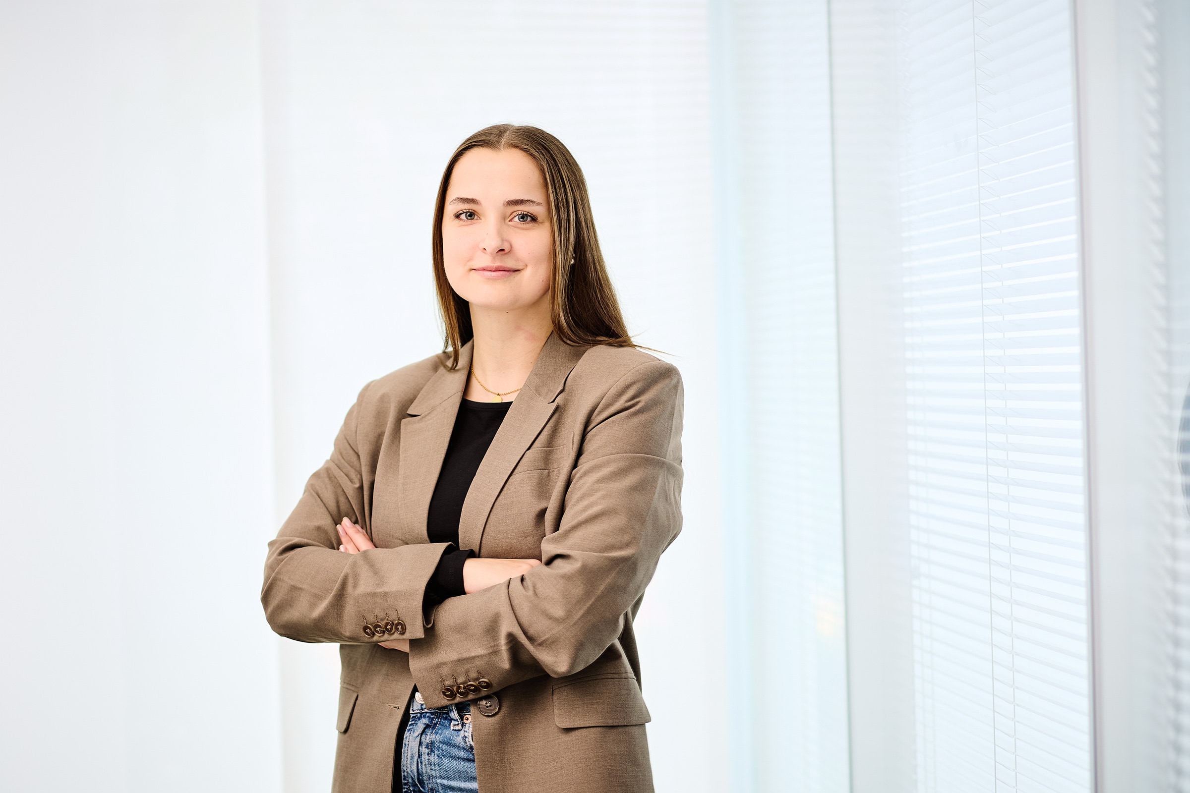 Katharina-Sophia  Schulz steht mit verschränkten Armen vor einem hellen Hintergrund und lächelt in die Kamera.