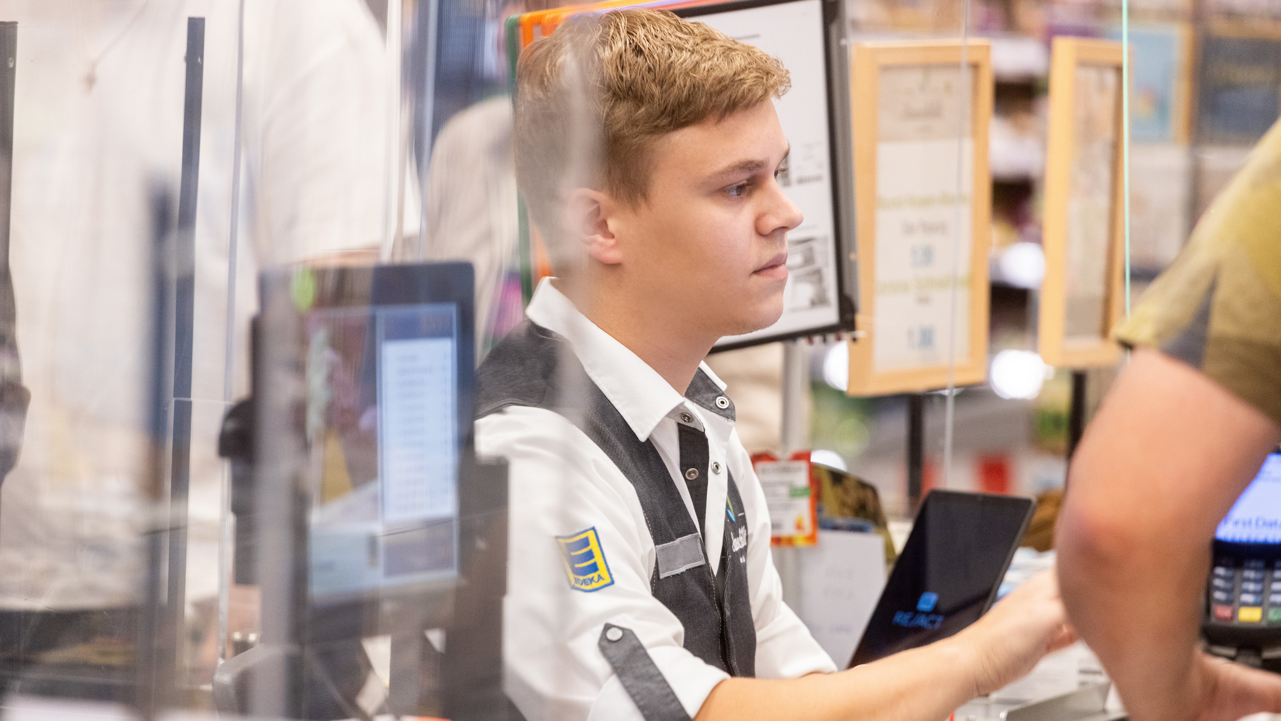 Ein junger Mann sitzt an der Kasse eines EDEKA-Einkaufsmarktes. Er trägt ein weißes Hemd und eine schwarze Weste.