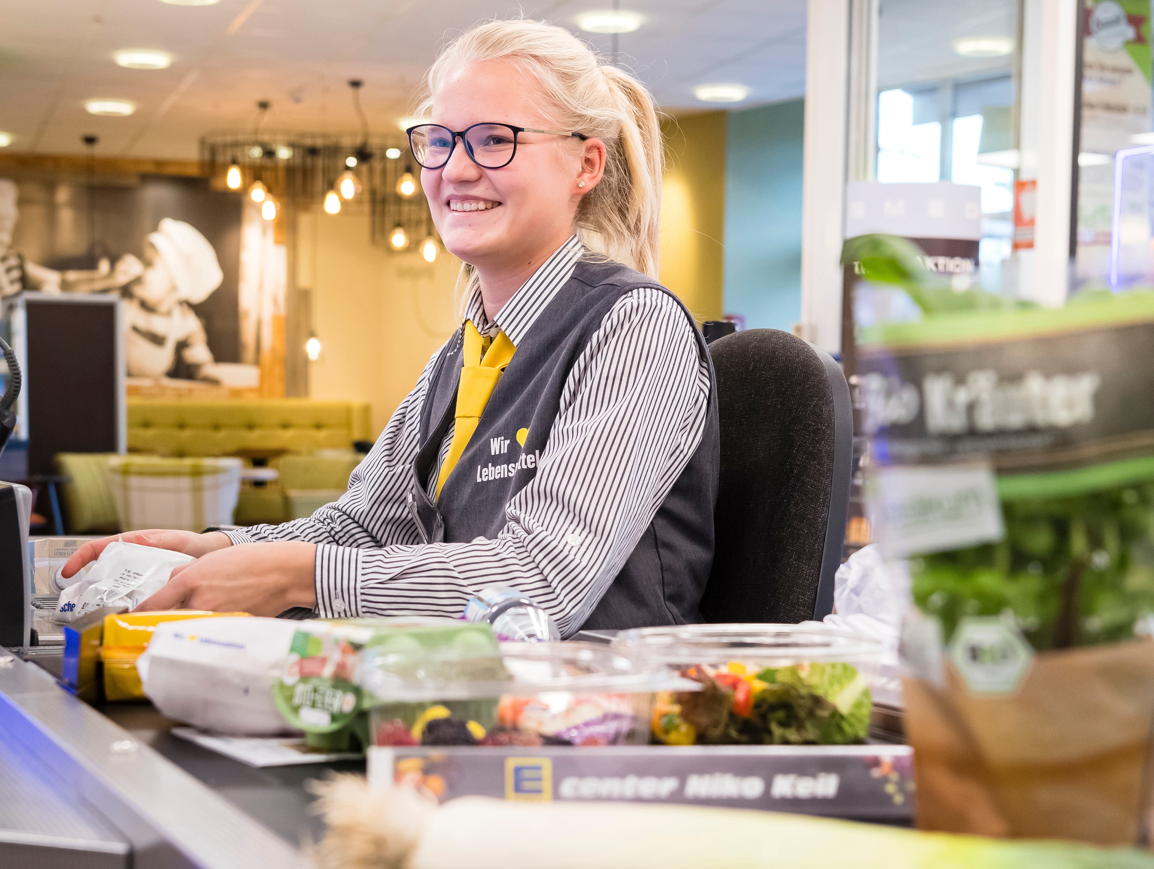Eine junge blonde Frau sitzt lachend an der Kasse eines EDEKA-Marktes.
