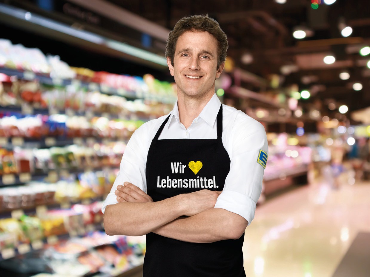 Ein Mann mit dunklen Haaren steht in einem EDEKA-Markt. Er trägt ein weißes EDEKA-Hemd und eine schwarze Schürze.