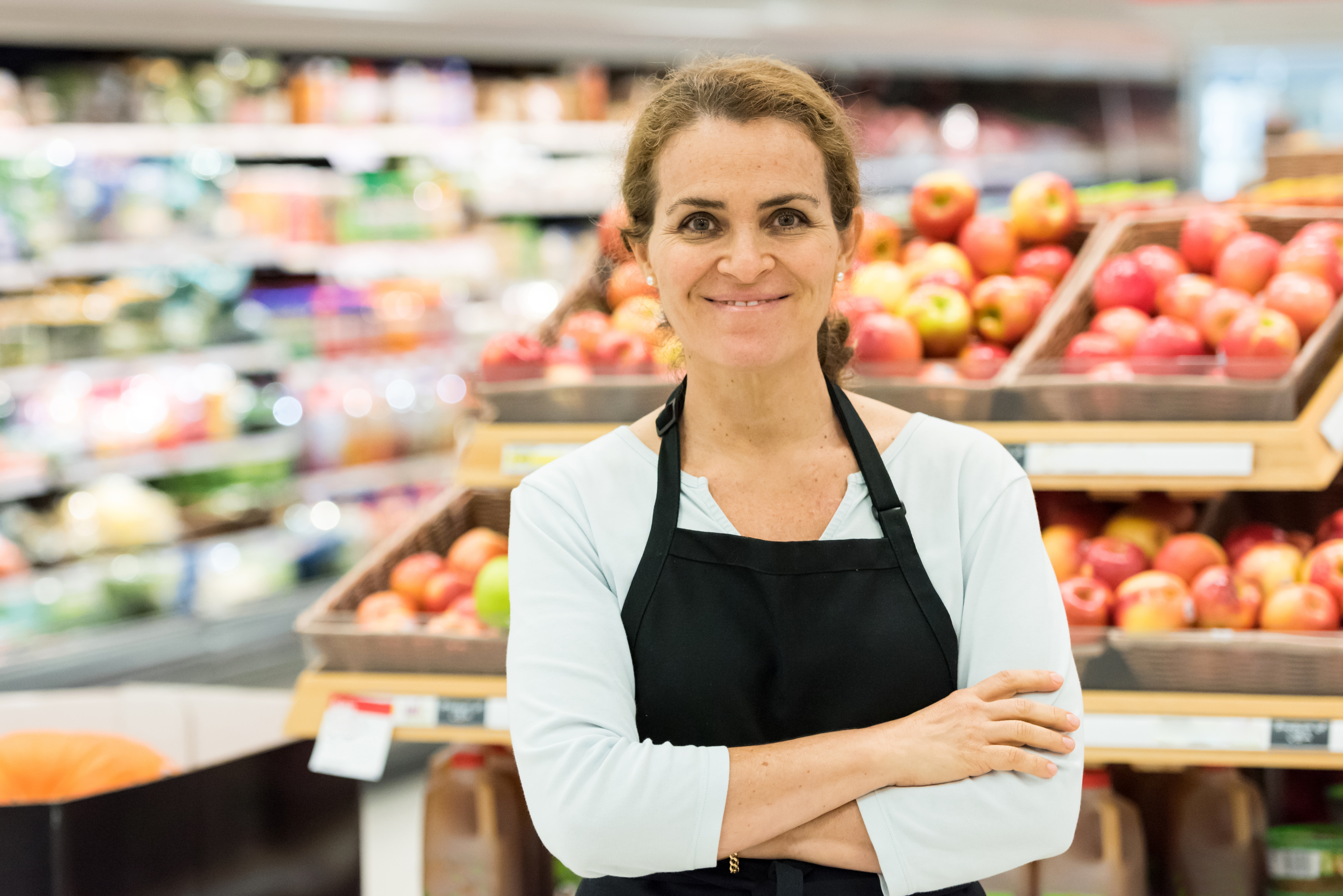 Eine Frau mit dunklen zurückgebundenen Haaren steht mit verschränkten Armen vor dem Apfelregal eines EDEKA-Marktes. 