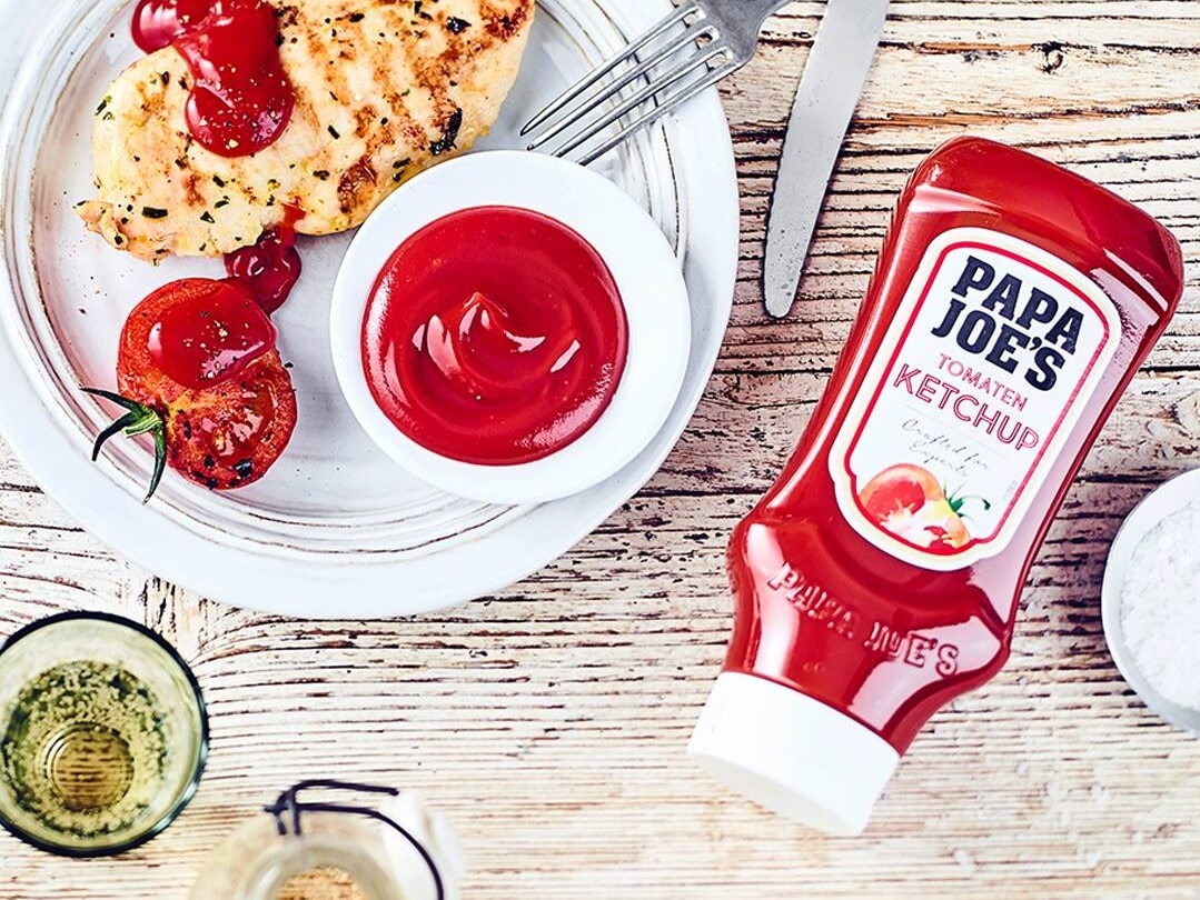 Eine Flasche Papa Joes Ketchup liegt auf einem Holztisch. Auf dem Teller daneben ein Stück Fleisch und eine halbe Tomate.