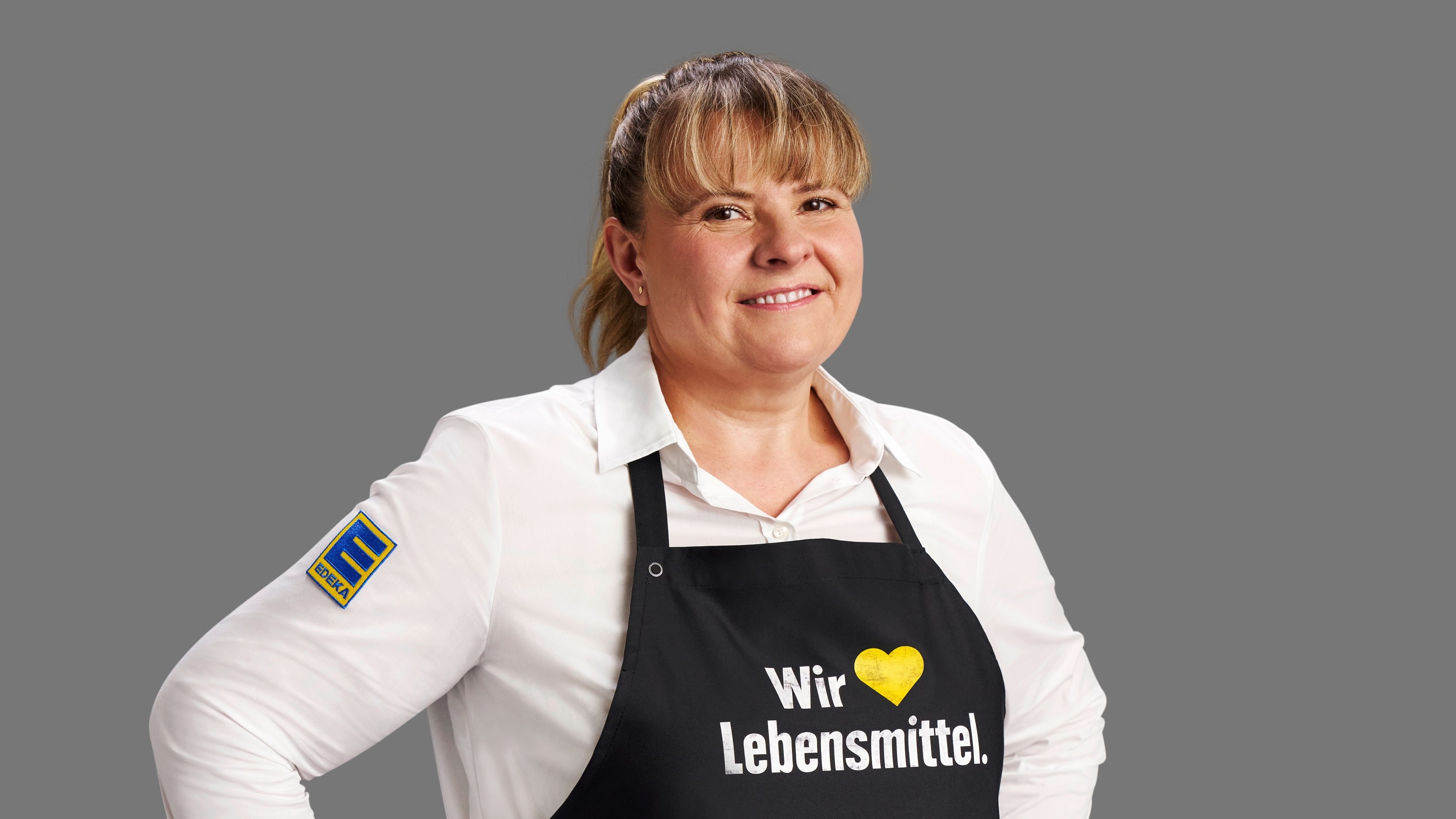 Karen Bich vor grauem Hintergrund. Die blonde Frau trägt ein weißes Hemd und eine schwarze Schürze mit Aufschrift.
