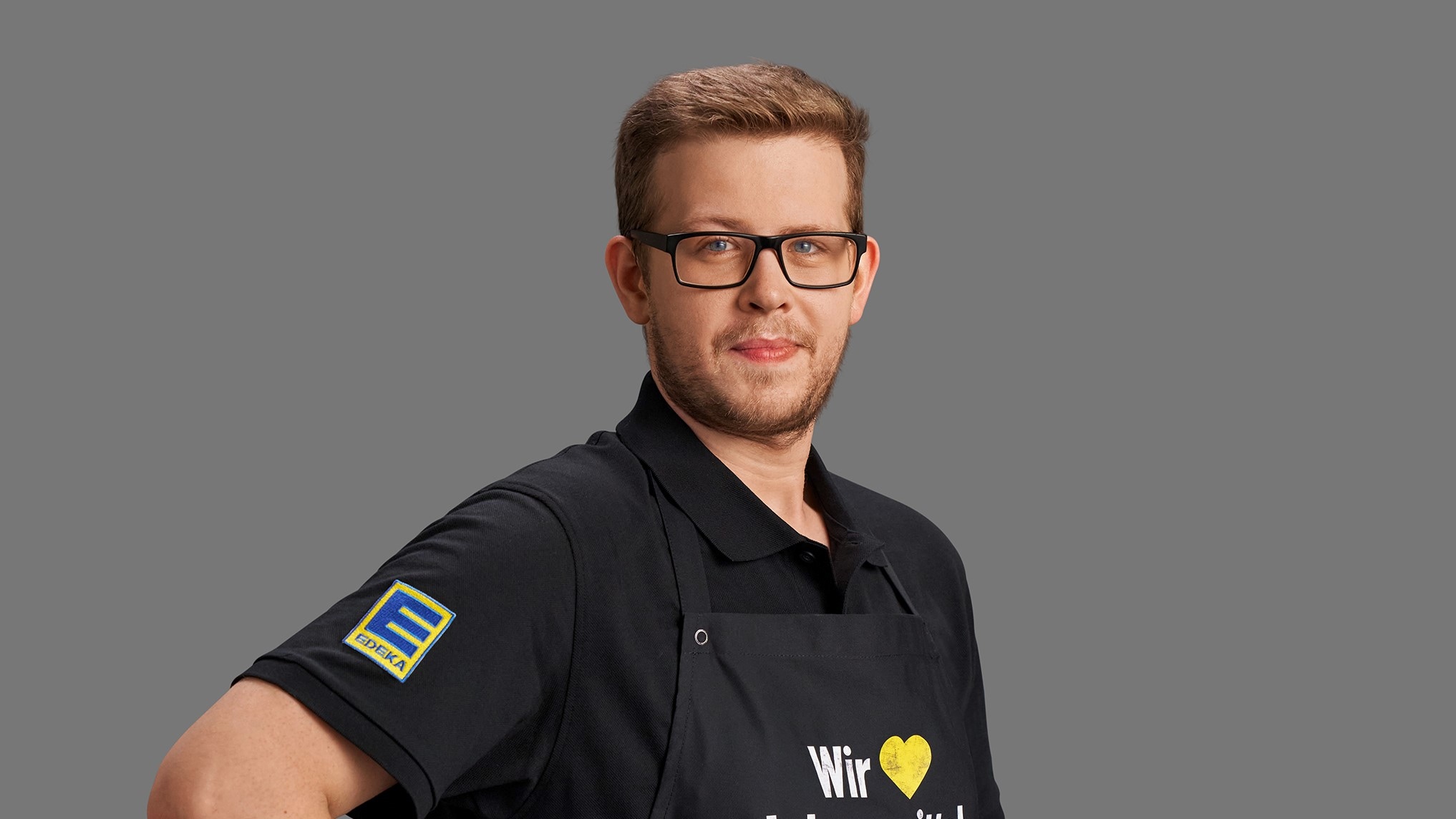 André Gruber vor grauem Hintergrund. Er trägt eine eckige Brille, ein schwarzes Polo-Shirt und eine schwarze Schürze.