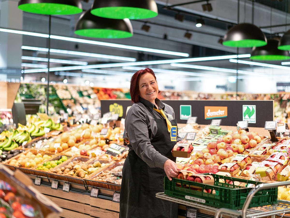 Eine Fachkraft mit EDEKA-Schurz steht lächelnd in der Obstabteilung und räumt Äpfel ein