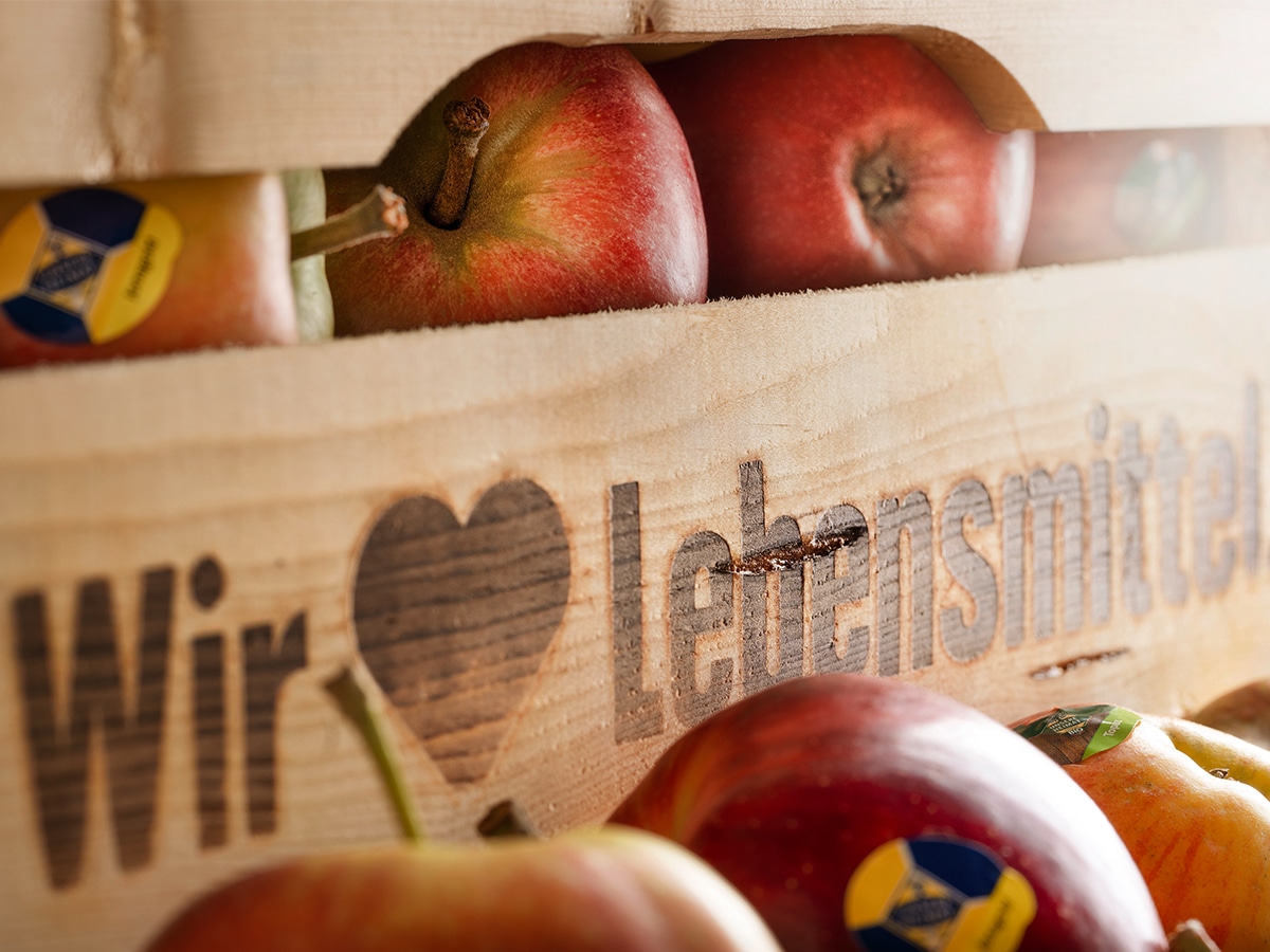 Bildausschnitt einer Holzkiste mit dem Aufschrift Wir ♥ Lebensmittel. Darin sind rote Äpfel mit Aufklebern "Unsere Heimat"
