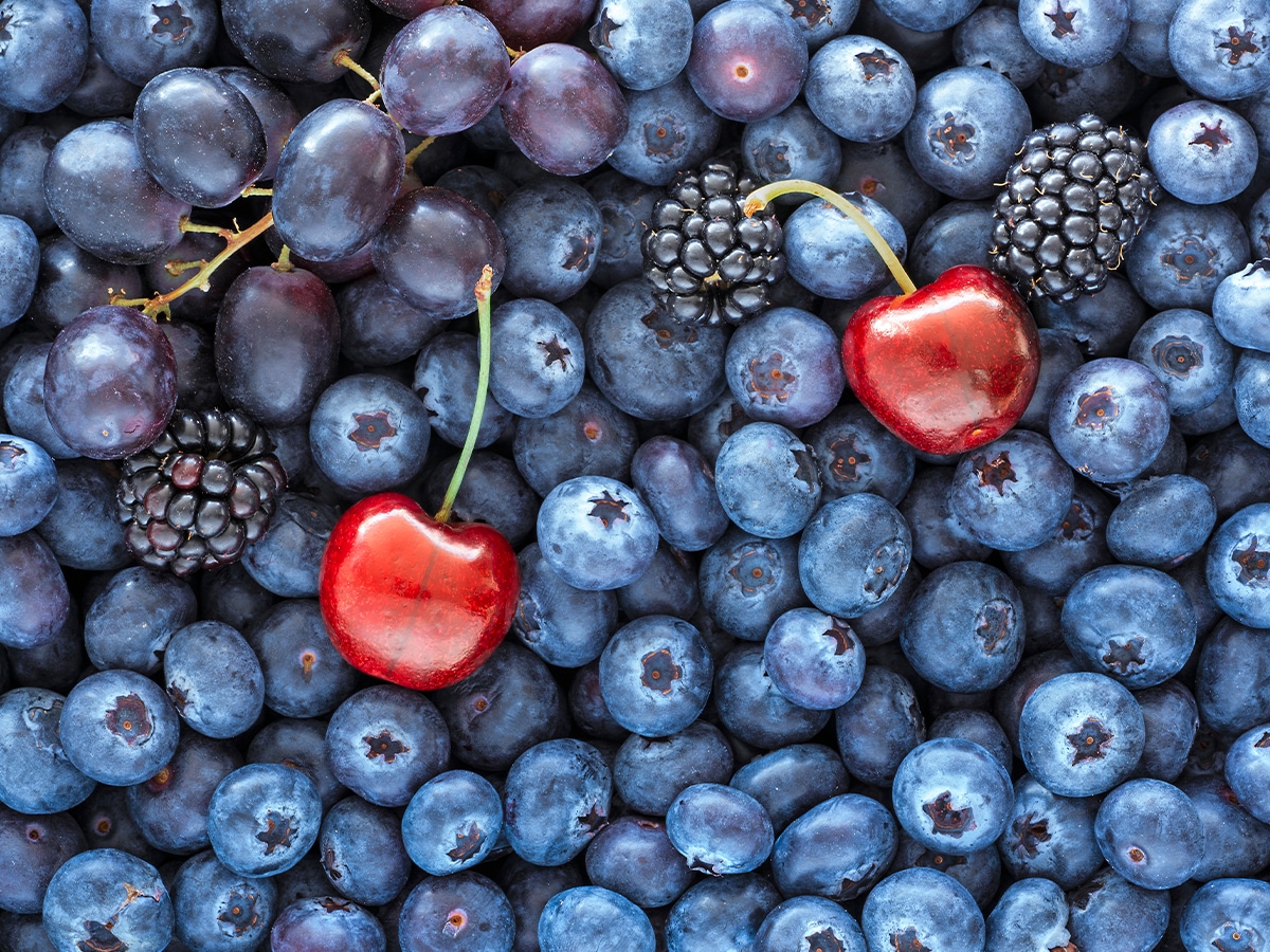Frische Blaubeeren als Untergrund, darauf liegen blaue Trauen, 3 Brombeeren und 2 rote Kirschen, die hervorstechen.