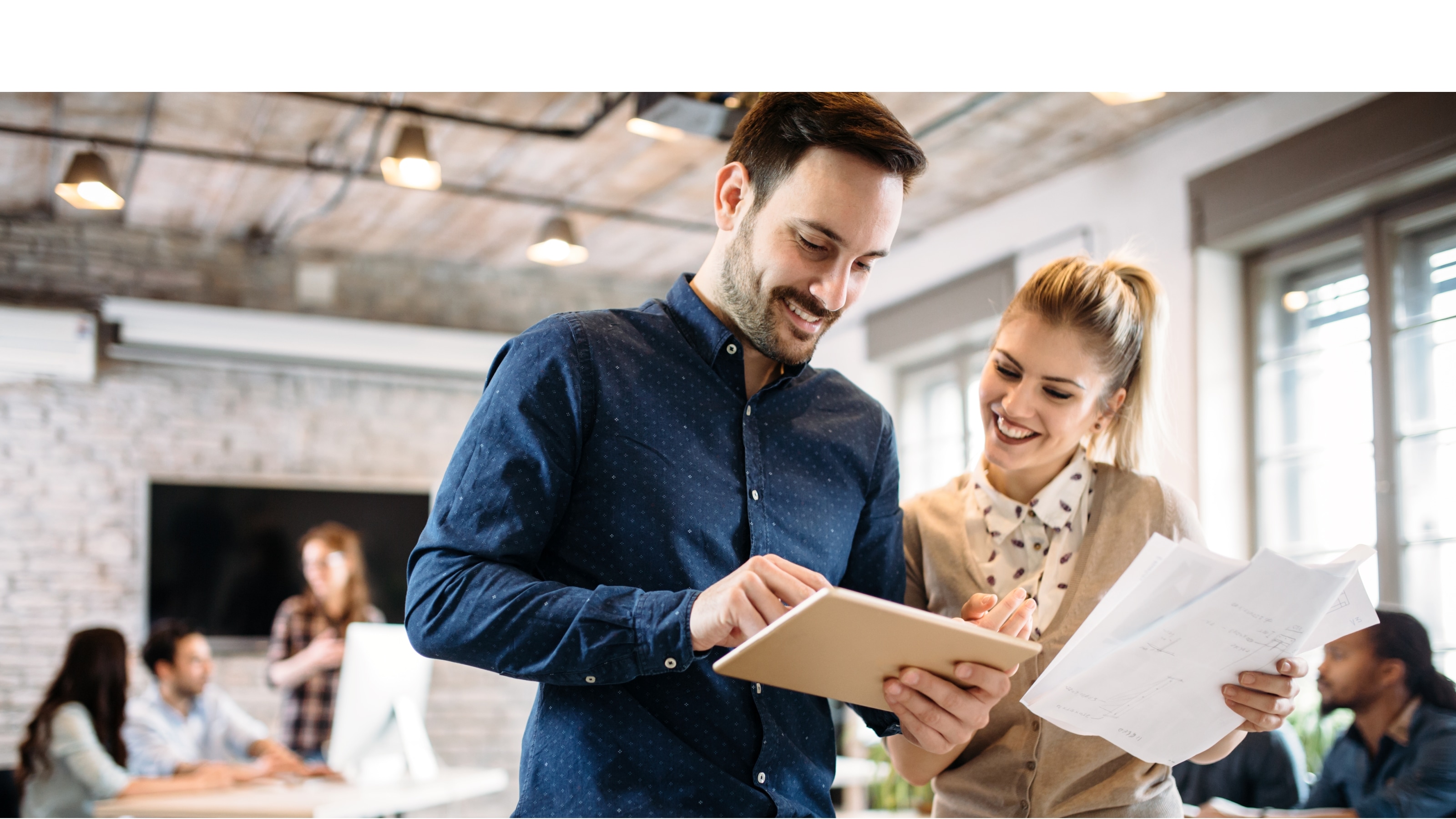 Taste your Job - Mann und Frau stehen im Büro zusammen und schauen mit Unterlagen in der Hand auf ein Tablet.