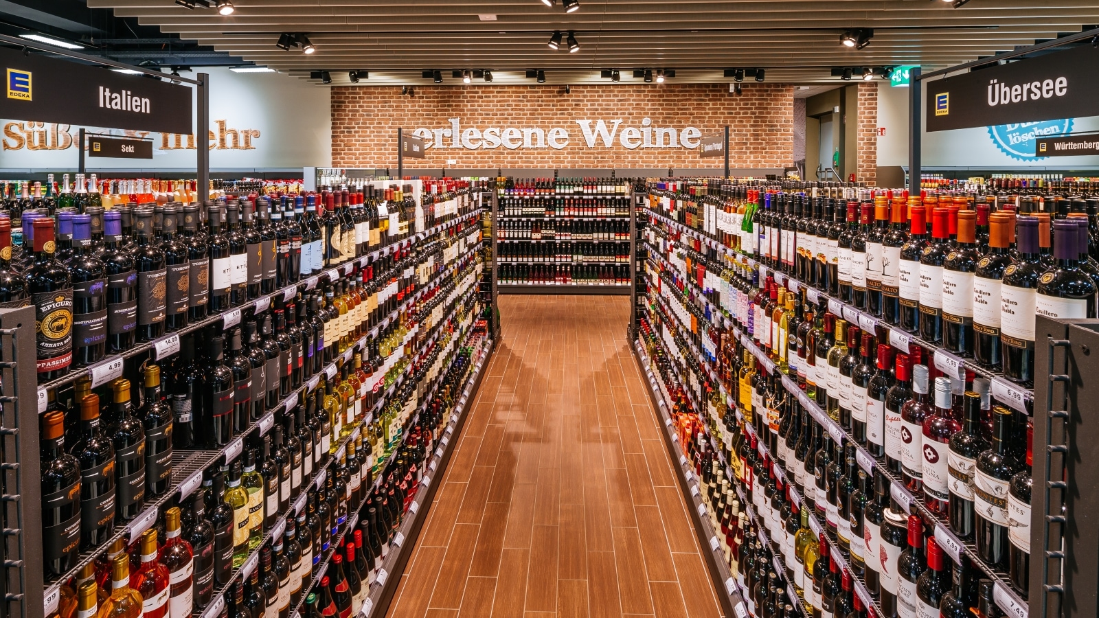 Weinabteilung: Regale mit Weinflaschen gefüllt, Blick nach hinten an der Wand. Über dem Regal quer steht "erlesene Weine".