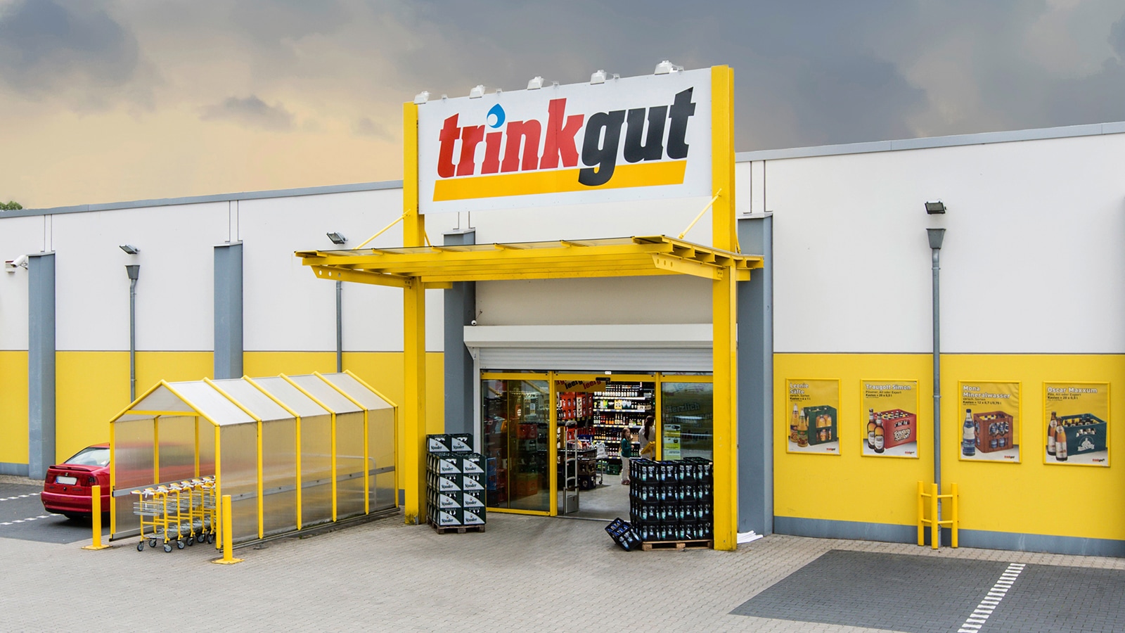 Außenansicht: Markteingang Trinkgut-Getränkemarkt, Getränkekisten daneben und großes Logo über dem überdachten Eingang.