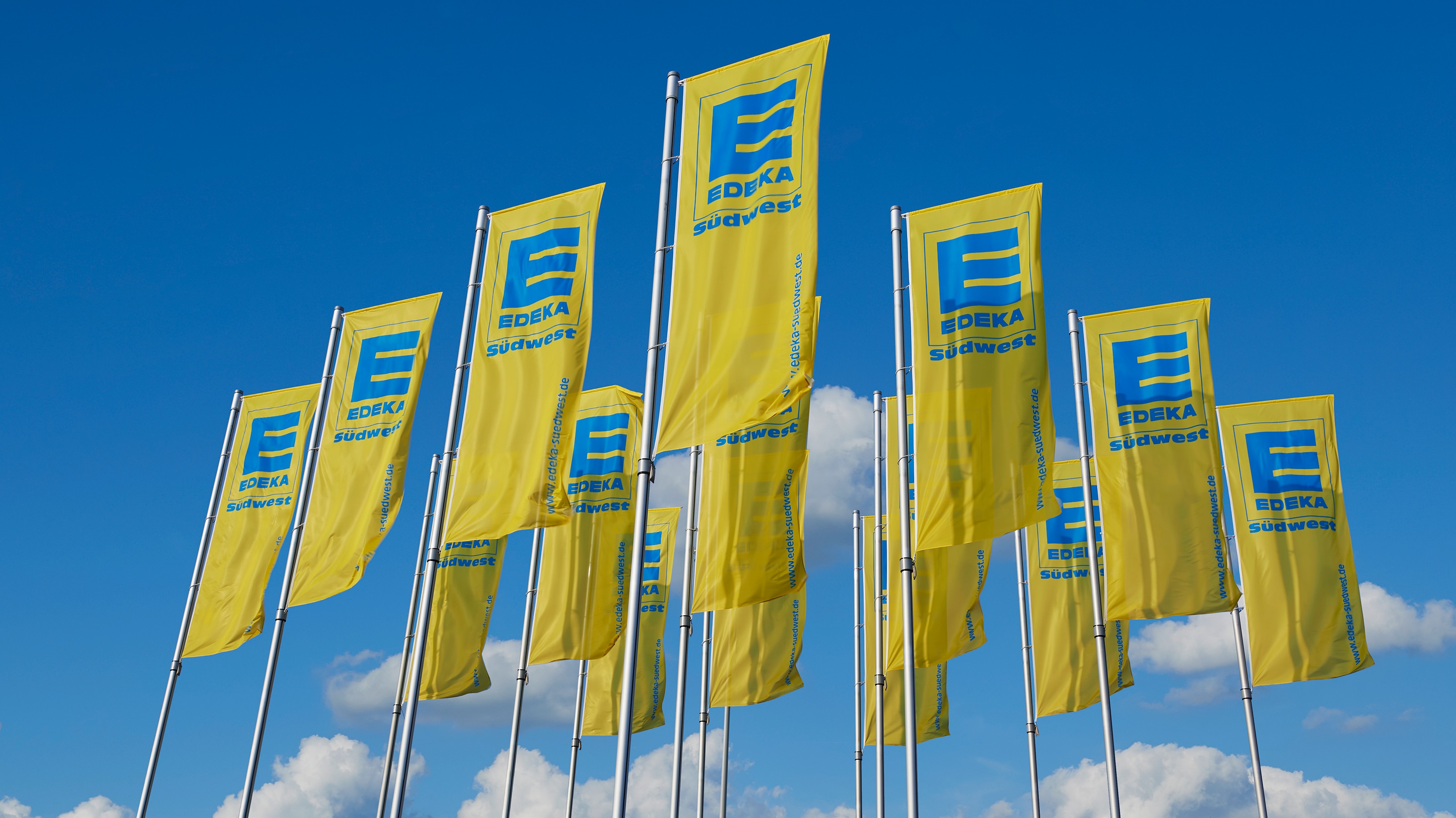 15 gelbe Fahnen mit der blauen Aufschrift EDEKA Südwest hängen an Masten und wehen im Wind, dahinter blauer Himmel.