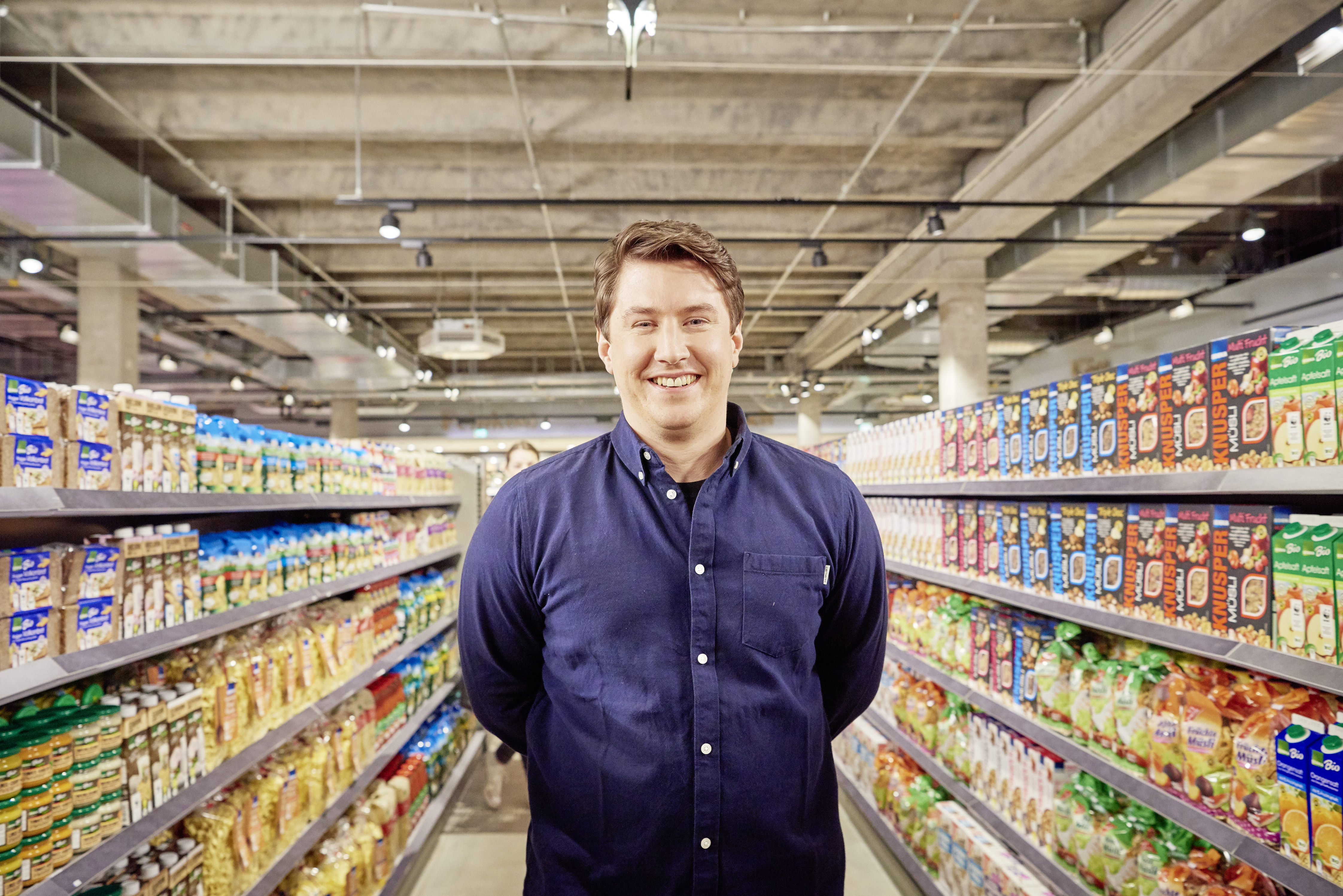    	 Ein lachender Mann steht im Gang eines Supermarktes mit gut sortierten Regalen zu seiner linken und rechten Seite.