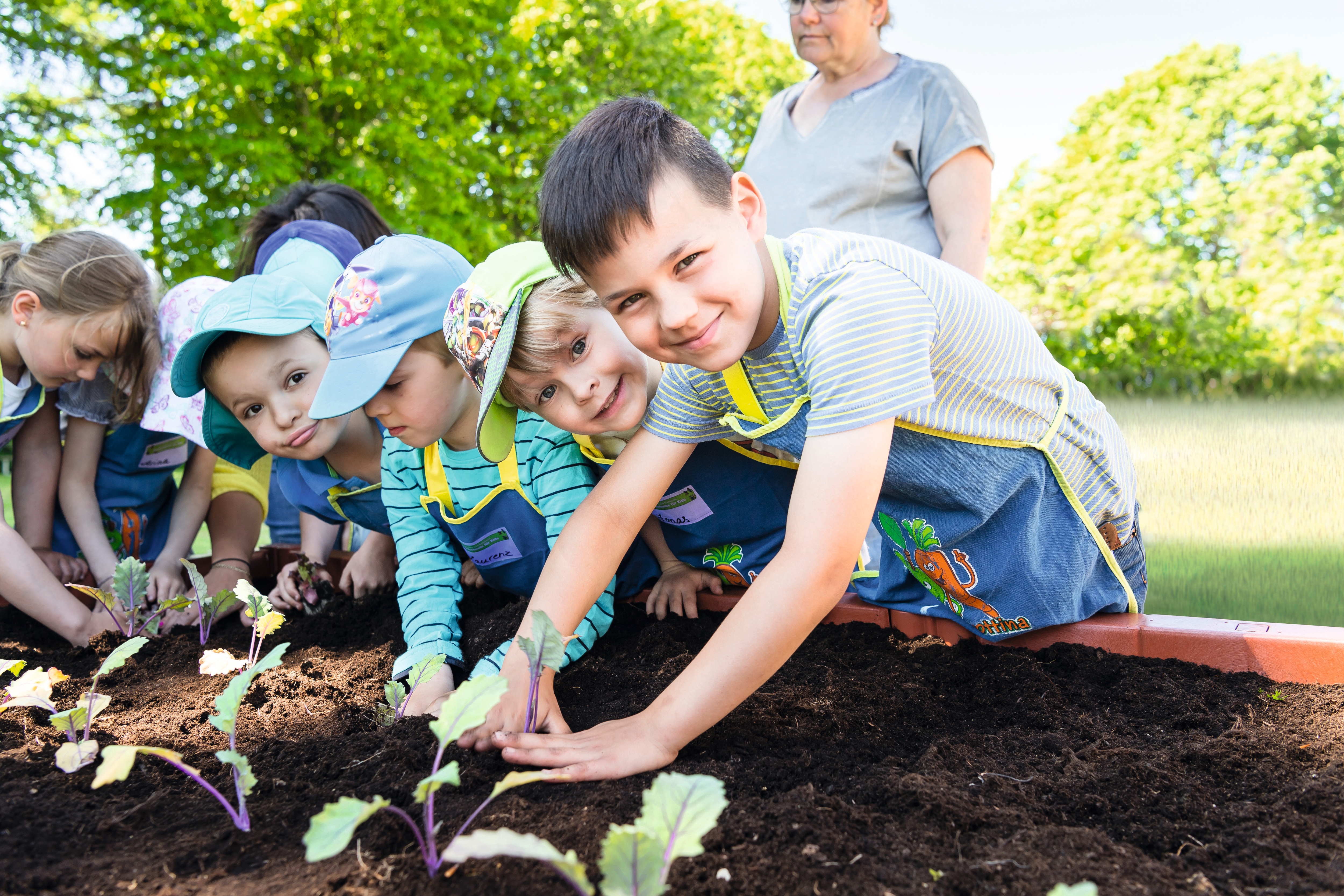 Mehrere Kleinkinder lehnen sich über ein Hochbeet. Sie tragen blau-gelbe Schürzen und graben mit ihren Händen.
