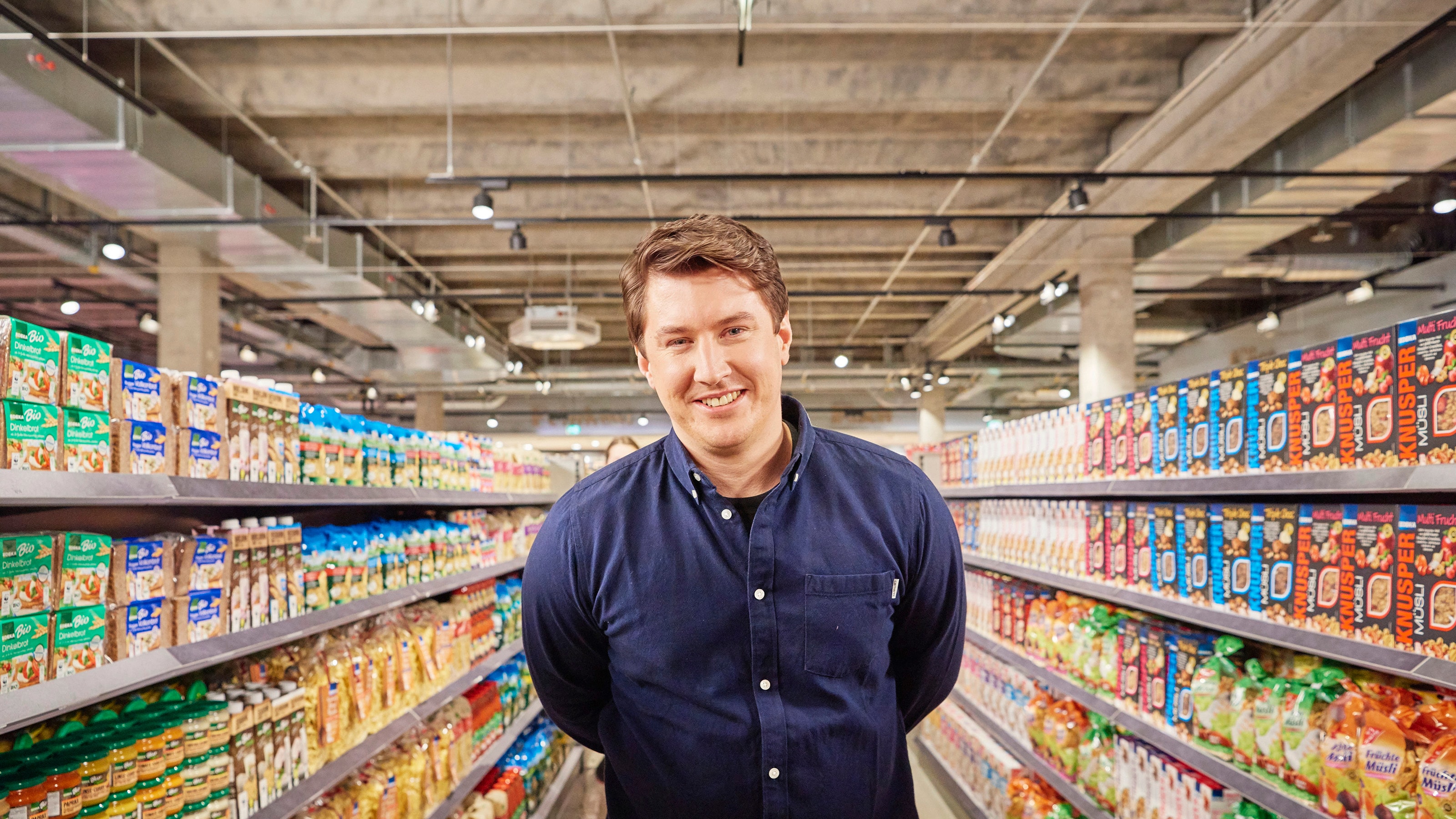 Ein lachender Mann steht im Gang eines Supermarktes mit gut sortierten Regalen zu seiner linken und rechten Seite.