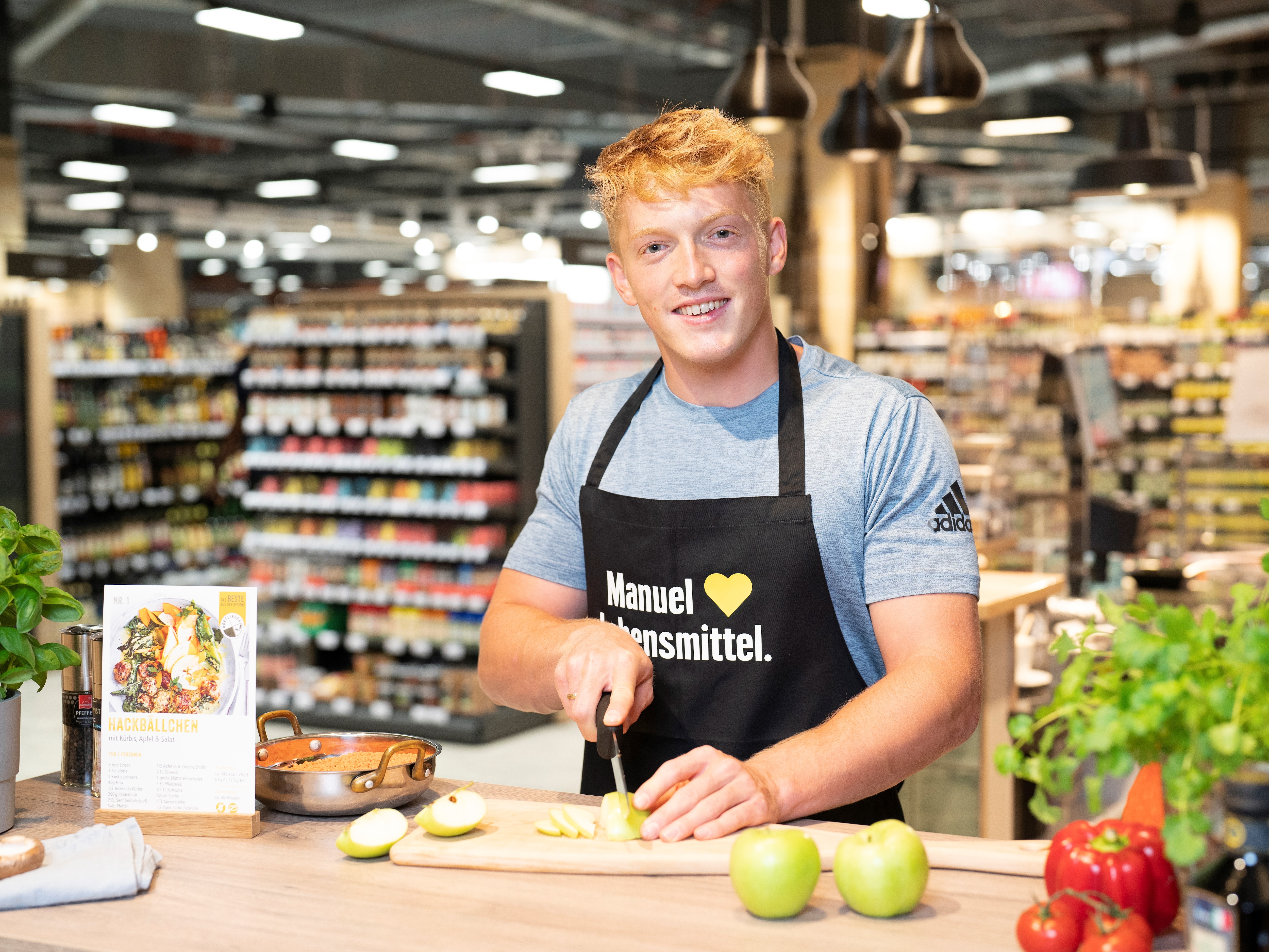 Ein Athlet steht hinter einem Tresen im EDEKA-Markt. Er schneidet grüne Äpfel und lächelt in die Kamera.