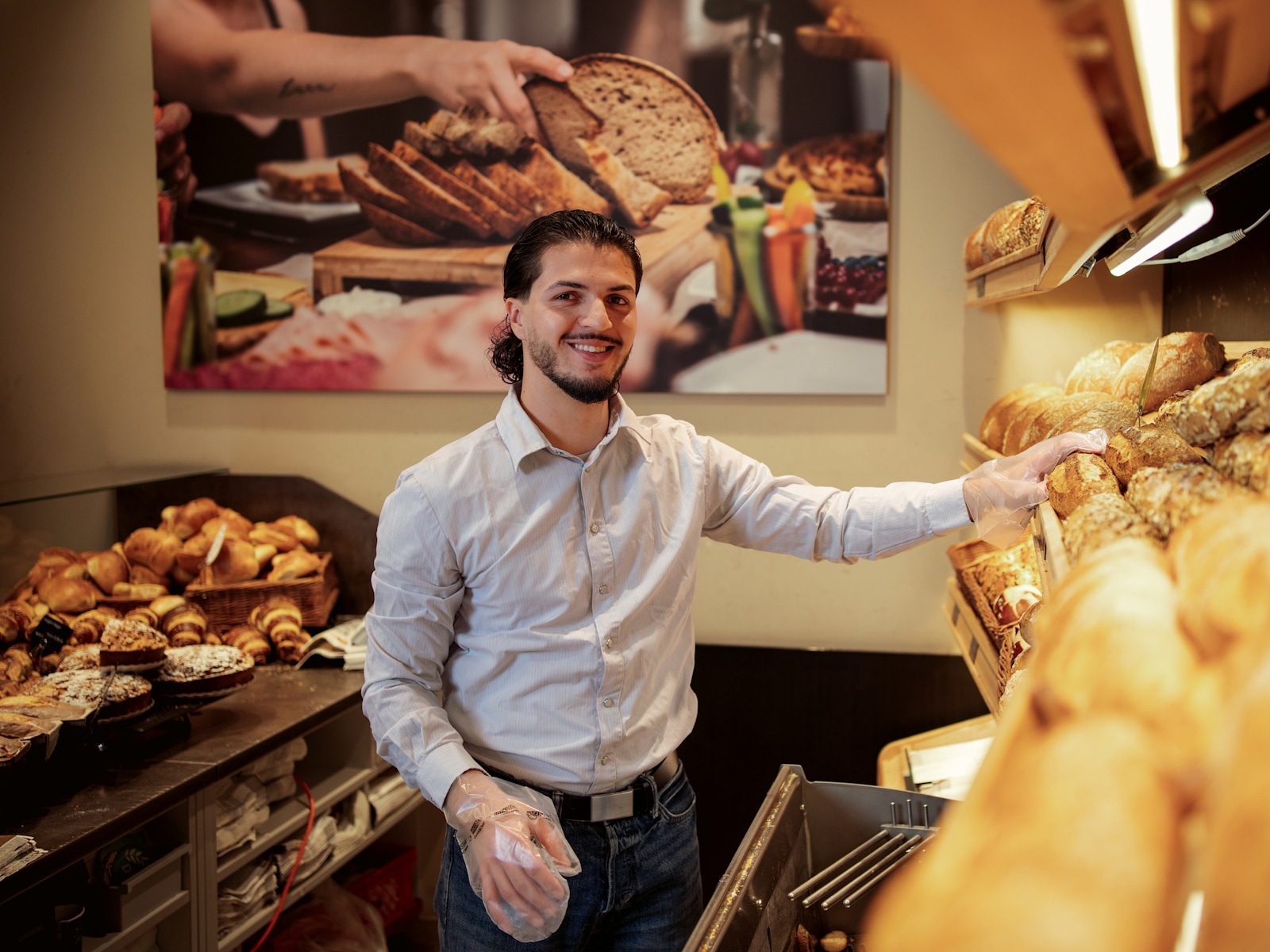 Ein junger Mann mit Plastikhandschuhen steht in der Backwarenabteilung hinter der Theke und greift nach einem Brot im Regal.
