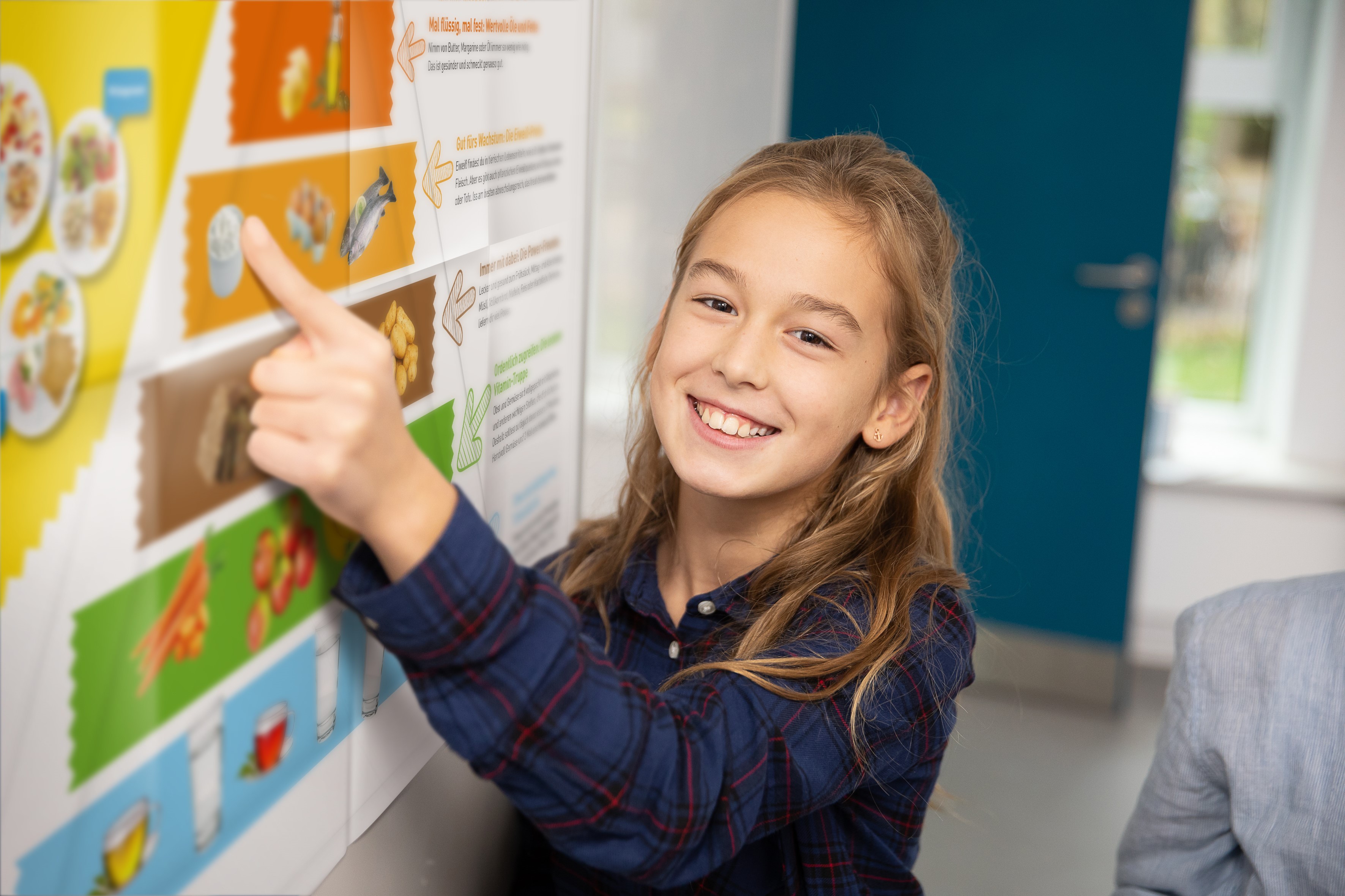 Ein Mädchen im Schulalter lacht und zeigt mit dem Zeigefinger auf ein großes Plakat mit der Ernährungspyramide..